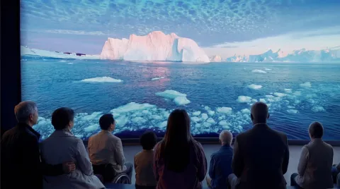 A group seated in a dark room watches a large screen showing an iceberg, sea ice, and calm ocean under clouds.