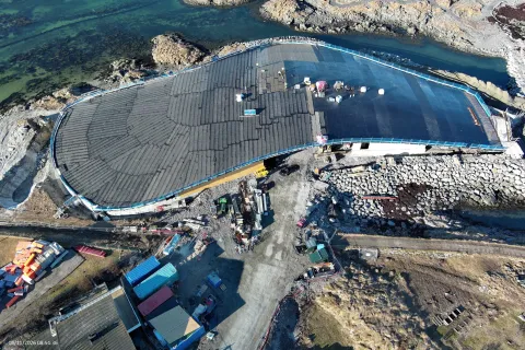 Aerial view of an oval roofed structure under construction beside rocky coastline, with machinery and materials below.