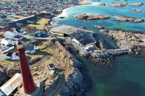 Aerial view of a red lighthouse beside a rocky shoreline, curved harbor building, and coastal village by clear blue sea.
