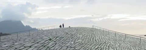 Two people stand on a curved stone-paved platform with railings, mountains on the left and seabirds in a cloudy sky.