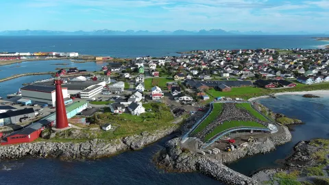 Aerial view of a seaside town with a tall red lighthouse on rocky shore, harbor buildings, and ocean beyond.