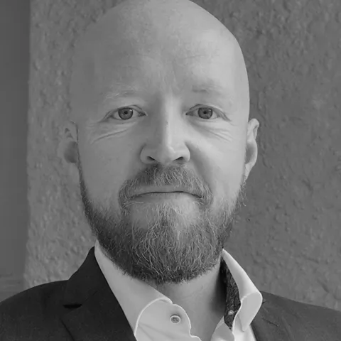Black-and-white head-and-shoulders portrait of a bald bearded man in a suit jacket and white shirt by a textured wall.