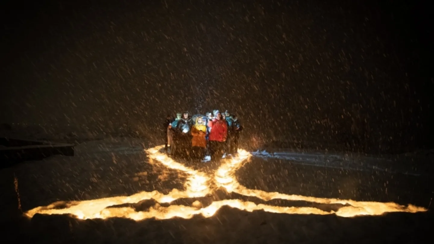 A group of people in winter jackets stands in falling snow at night, surrounded by glowing lines of fire on the ground.