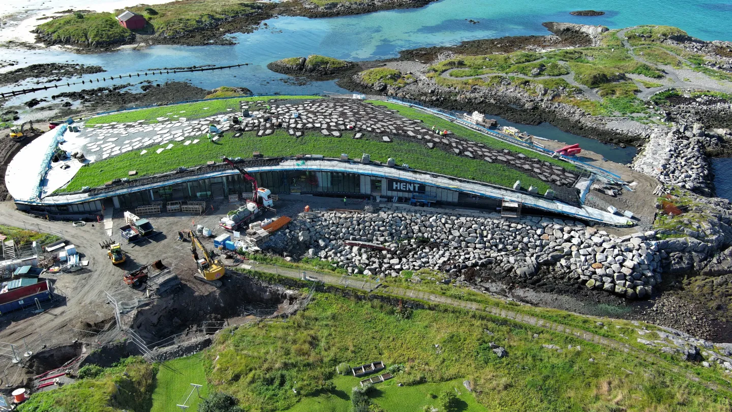 Aerial view of a curved coastal building under construction with a green roof, rock barrier, and blue water bays.