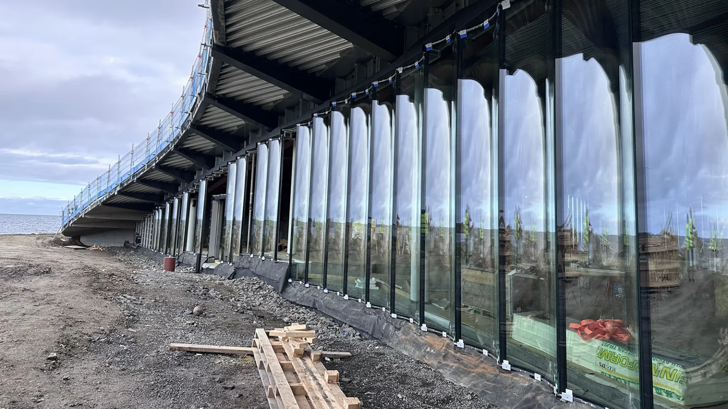 Curved structure with tall glass panels under a metal deck, beside a rocky shoreline with construction materials.