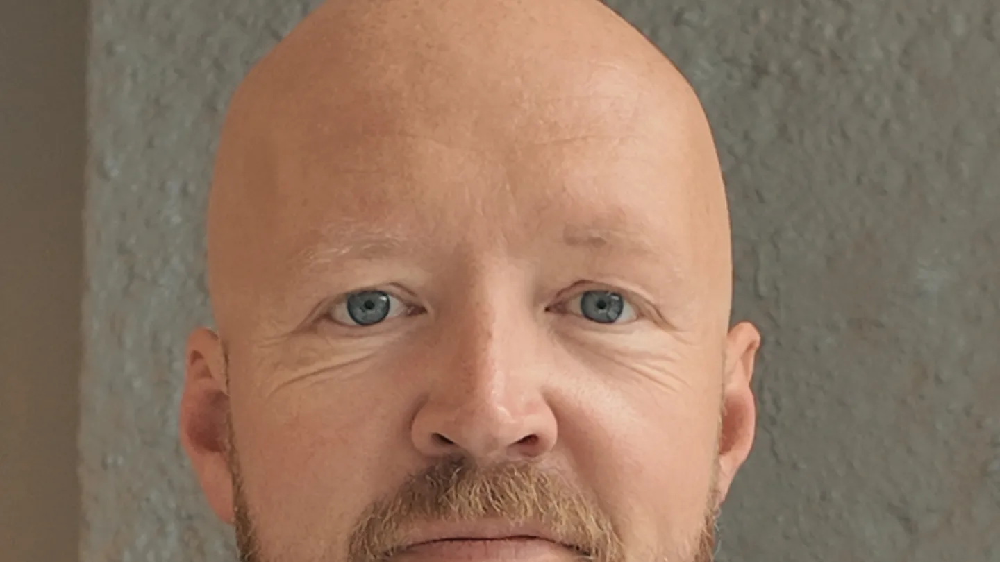 Close-up portrait of a bald bearded man in a dark suit and white shirt, standing against a gray wall.