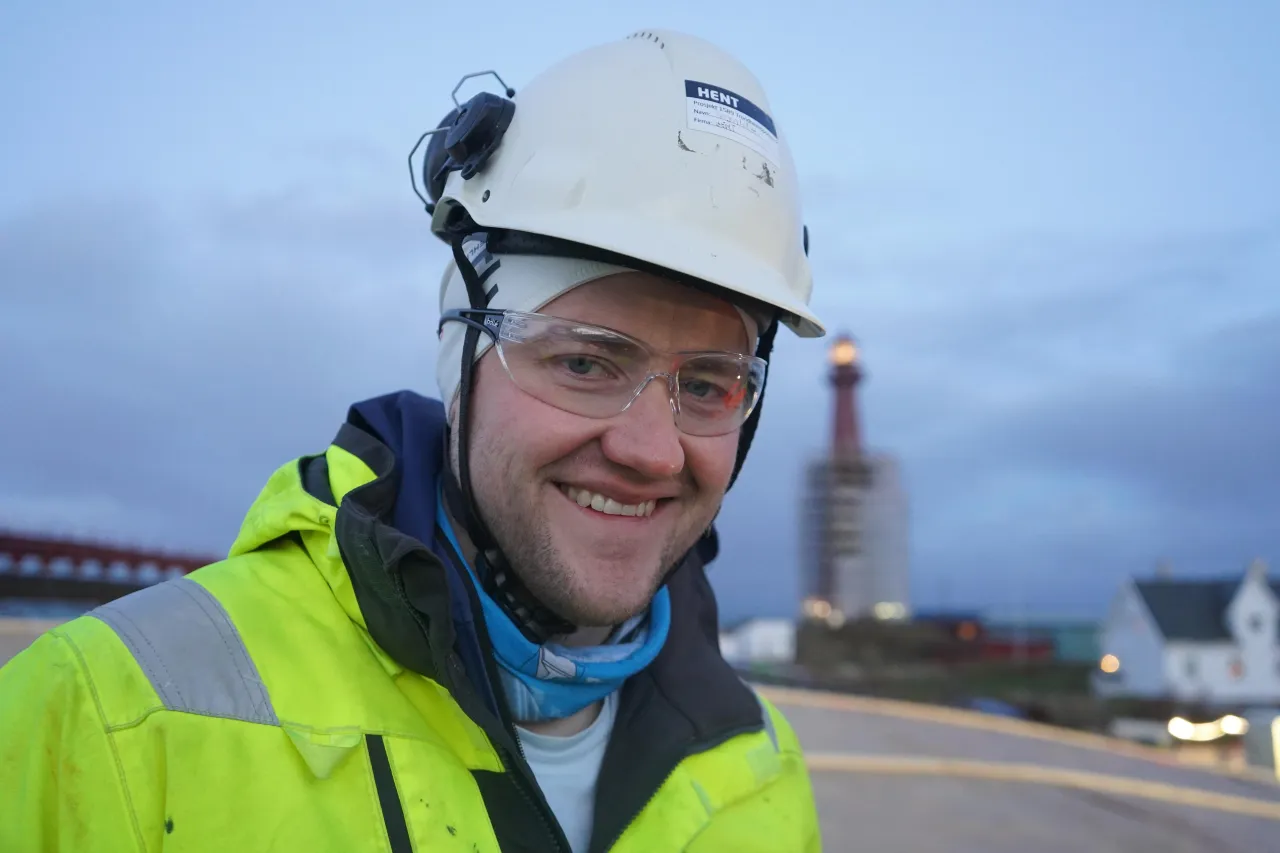 Person in a high-visibility jacket and white hard hat stands outdoors near a road with a tower in the distance.