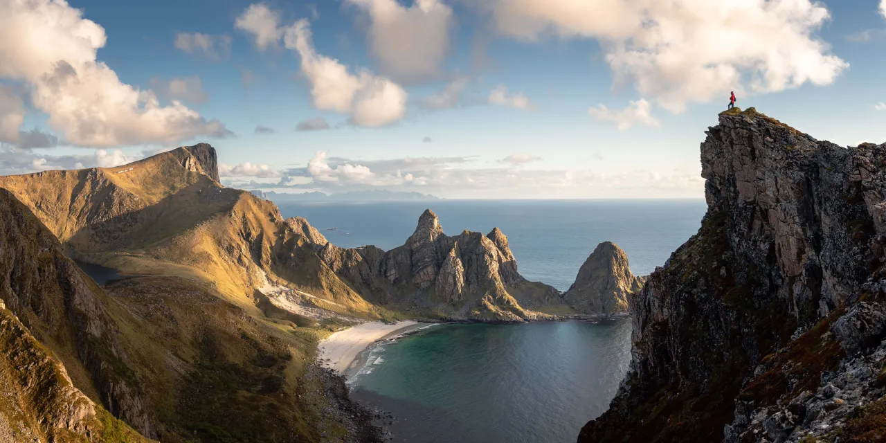 Wide view of rugged coastal mountains and a crescent beach below, with a lone hiker standing atop a rocky cliff.