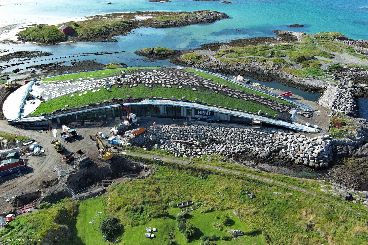 Aerial view of a curved building with a green roof under construction beside rocky shoreline and turquoise water.