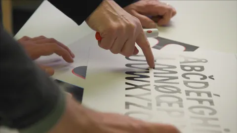 Close-up of hands pointing at printed letterforms on paper during a design review at a desk with markers nearby.