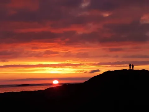 Two people stand silhouetted on a rocky ridge, overlooking the ocean as the sun sets under red-orange clouds.