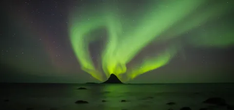 Green aurora curtains swirl across a starry night sky above calm water and a dark island silhouette on the horizon.