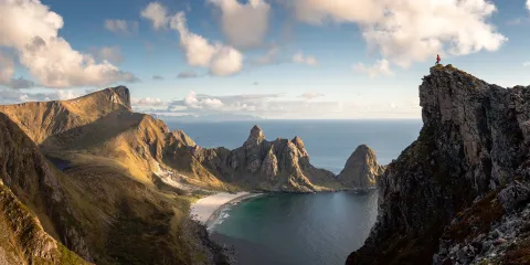Wide view of rugged coastal mountains and a crescent beach below, with a lone hiker standing atop a rocky cliff.