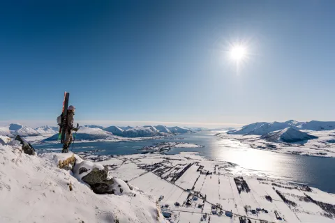 A skier with skis on their back stands on a snowy ridge above a fjord and mountains under a bright sun.