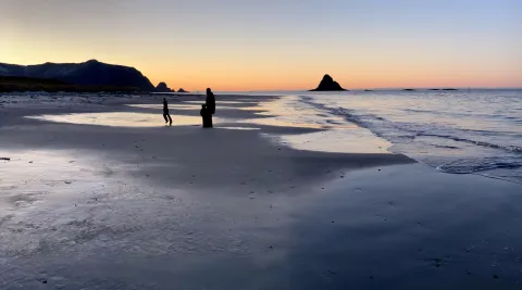 Two silhouetted people stand on wet sand at sunset, with gentle waves, distant mountains, and a sea stack offshore.