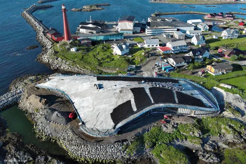 Aerial view of a coastal harbor with a red lighthouse, small village buildings, breakwaters, and a large slab under construction.