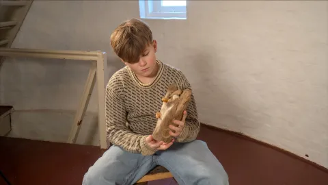 A seated person in a patterned sweater holds wooden clogs indoors near a stair railing and a small window.